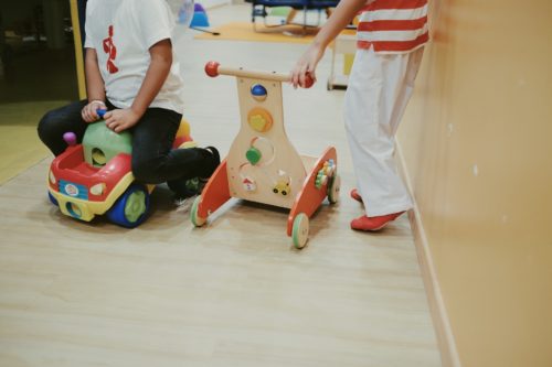 Two children are joyfully playing indoors at the daycare. One is sitting on a colorful ride-on toy car, while the other stands beside a wooden push toy with wheels. They are near a light brown wall on a wooden floor, giggling in their cozy corner of fun and adventure.