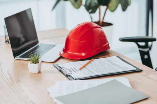 A desk with a laptop, a red hard hat, a clipboard with papers and a pencil, a small potted plant, and an office chair.