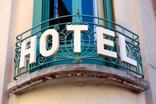 A hotel sign with large white letters is mounted on a green wrought iron balcony railing in front of a light-colored building with a window and green shutters.