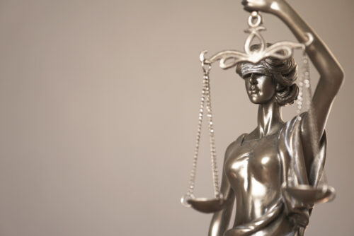 A close-up of a metallic Lady Justice statue holding balanced scales, with a blindfold over her eyes, against a plain, light background.